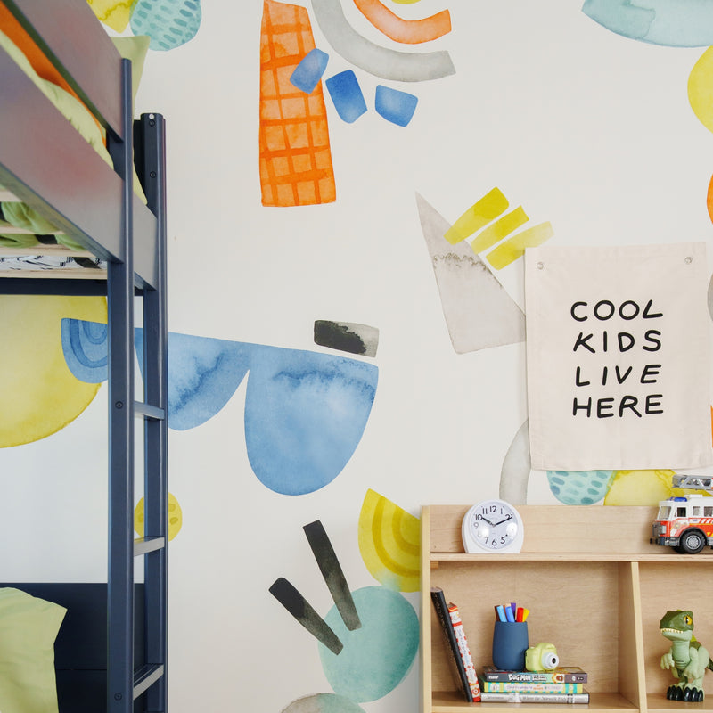 Children's bedroom decor: navy bunk bed, colorful abstract wall mural, wooden cubby shelf with clock, books, toy dinosaur and canvas sign reading "COOL KIDS LIVE HERE