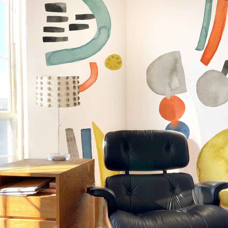 Modern home office corner with black leather lounge chair, wooden side table and textured pom-pom table lamp in front of a colorful abstract watercolor wall mural lit by natural window light