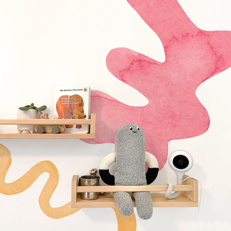 Nursery wall with pink watercolor mural and light wood floating shelves displaying the book "Brown Bear, Brown Bear, What Do You See?", a small succulent in a white planter, wooden toy car, gray knitted doll and white baby monitor — modern kids room decor and wooden toy shelf styling