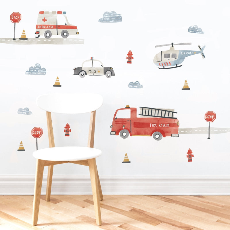 Kids room nursery wall decal in watercolor style featuring emergency vehicles — red fire rescue truck, police car, ambulance and air care helicopter with stop signs and traffic cones on a white wall behind a wooden chair