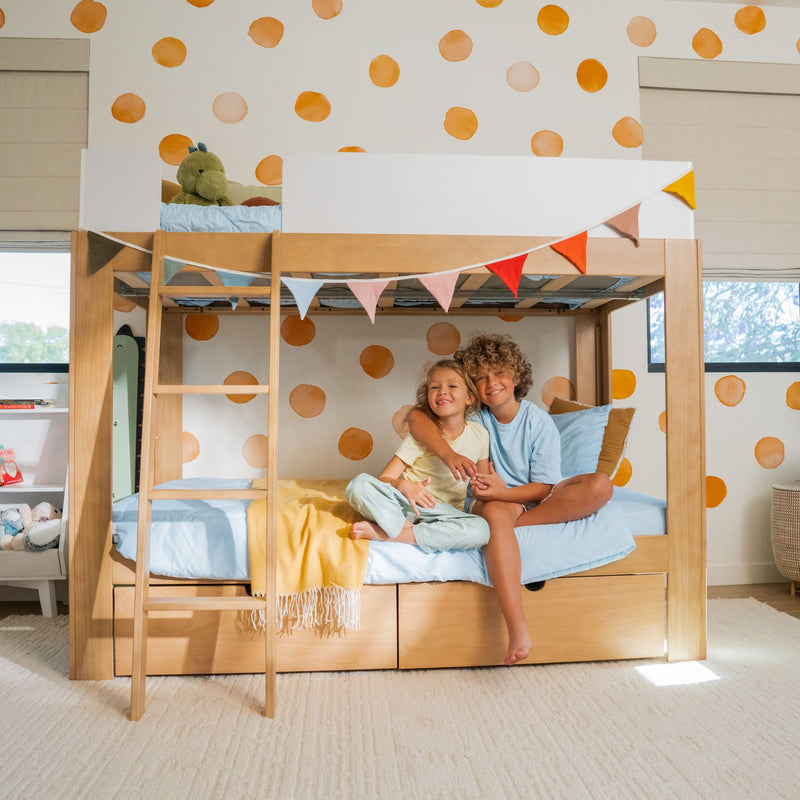 Two smiling children sitting on a wooden bunk bed in a modern kids' bedroom with orange polka dot wall, pastel bedding, colorful bunting and a plush dinosaur on the top bunk