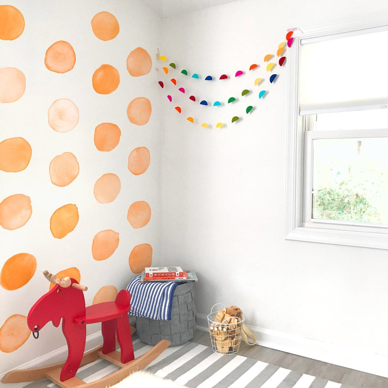 Bright modern nursery corner with orange watercolor polka dot accent wall, rainbow paper garland, red wooden rocking horse, gray storage basket with striped blanket and wire basket of wooden toys by a sunny window on a striped rug