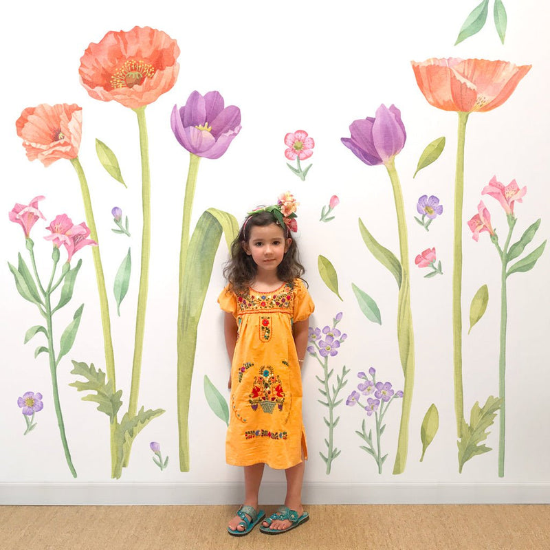 Little girl in a yellow embroidered dress and teal sandals standing in front of a large watercolor floral wall mural, children’s fashion portrait