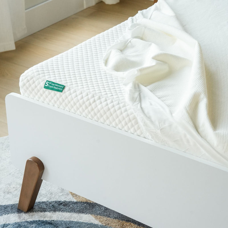 Memory foam mattress with white textured surface and sheet on a modern wooden bed frame in a bright bedroom setting.