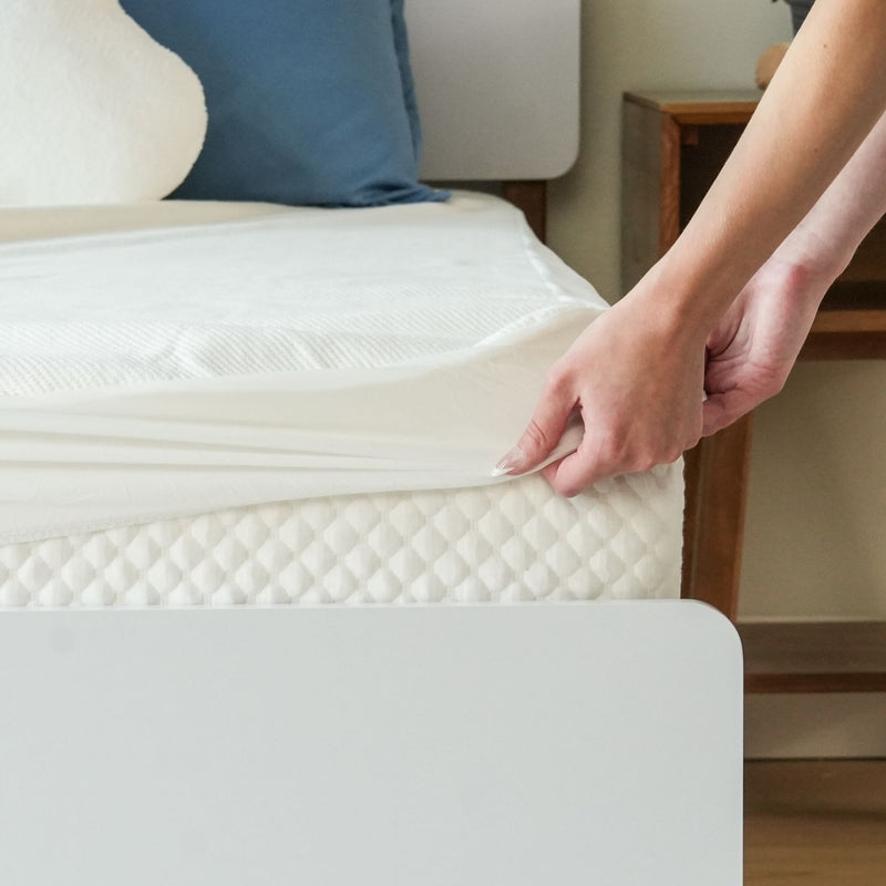 Person putting on a fitted sheet over a mattress with blue pillows in a modern bedroom setting.