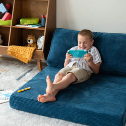 Smiling boy playing a handheld video game console on a blue sofa in a bright kids' playroom with toys on a wooden shelf