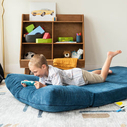 Child lying on a blue corduroy kids floor mattress playing a handheld game in a modern playroom with wooden toy shelf and stuffed animals