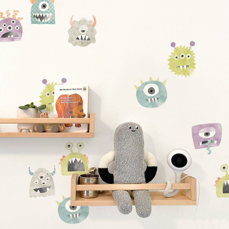 Nursery wall with colorful monster decals, wooden floating shelves holding knit doll, "Brown Bear" book, plant and baby monitor camera