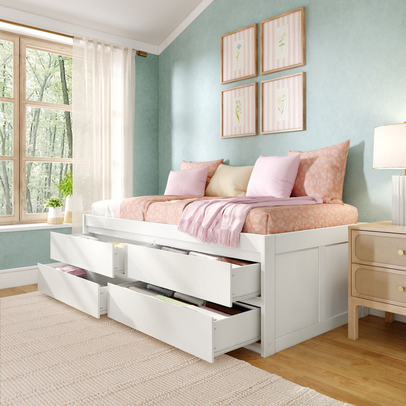 White space-saving storage bed with multiple open under-bed drawers, pink floral bedding and pastel pillows, mint-green accent wall with framed botanical prints, large wood-frame window and bedside lamp in a bright modern bedroom