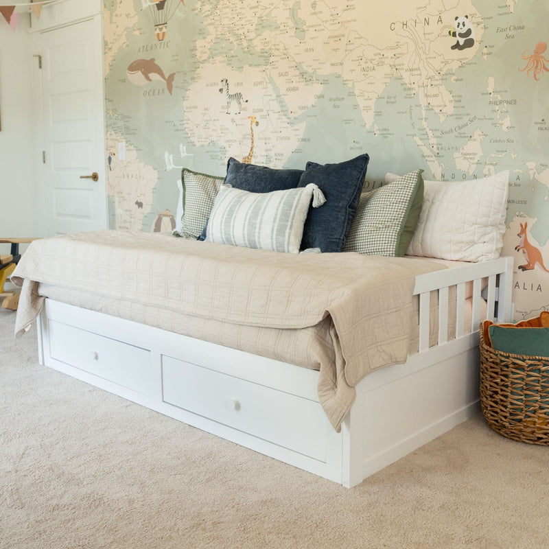 White daybed with storage drawers, neutral beige quilt and layered pillows against a pastel world map wallpaper in a kids' bedroom