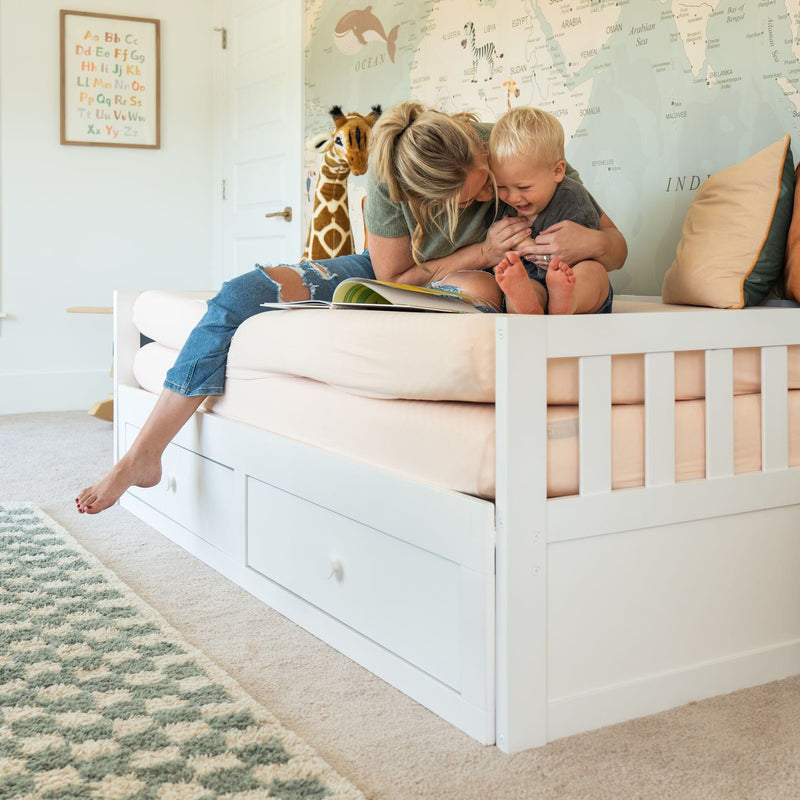 Mother reading and laughing with toddler on a white toddler bed with under-bed storage in a cozy nursery with world map mural and alphabet poster