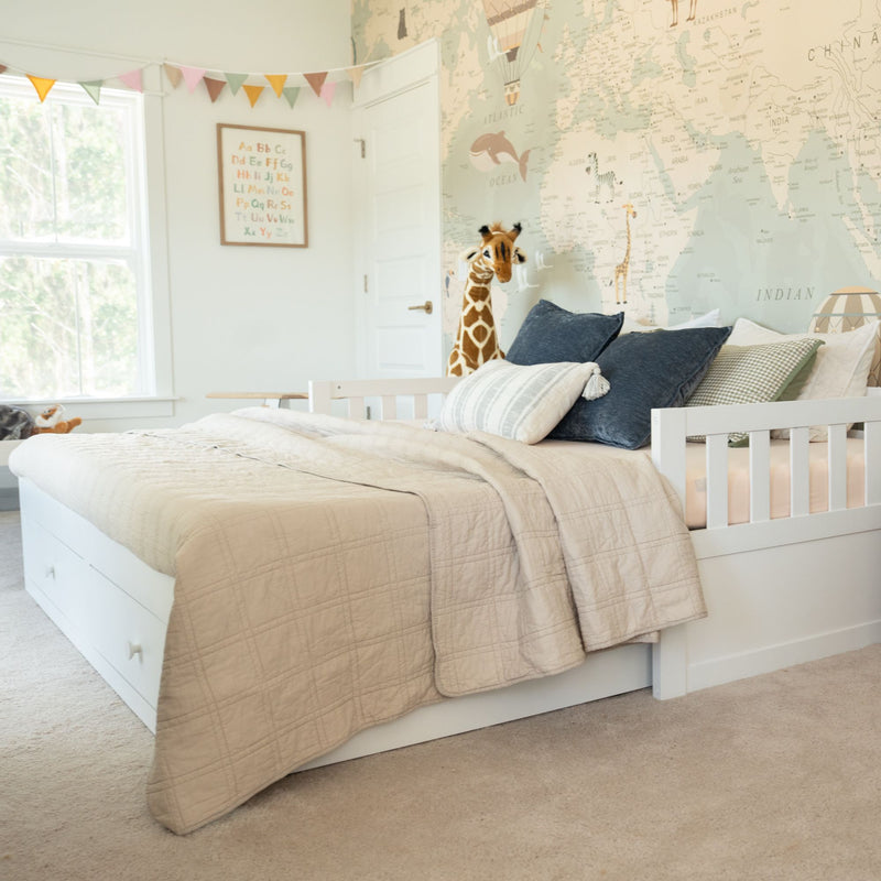 Cozy neutral kids bedroom with white daybed and storage, beige quilted bedding, navy pillows, world map mural and stuffed giraffe