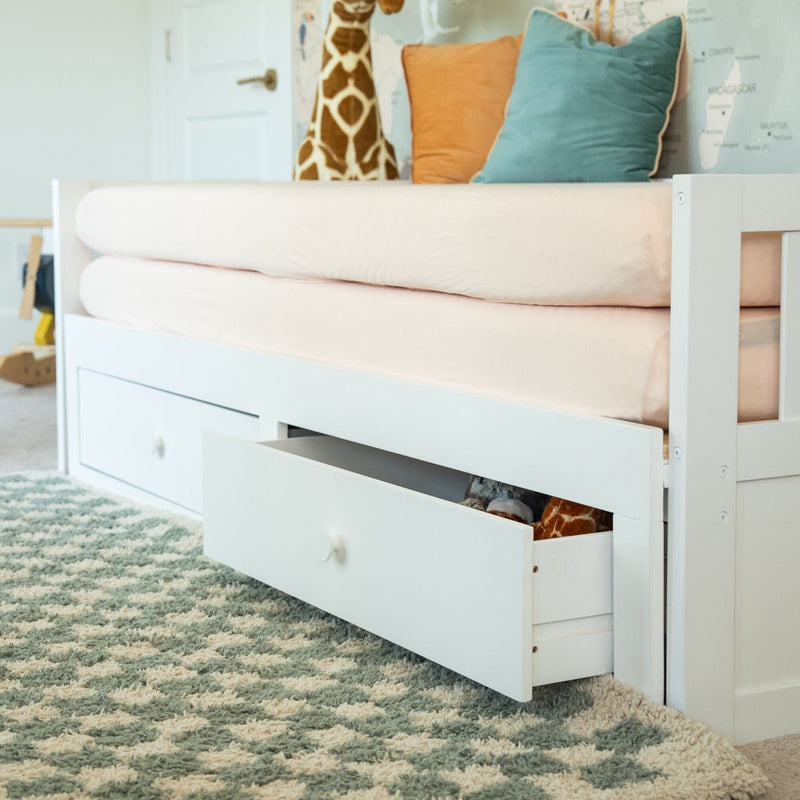 White wooden kids' daybed with pull-out storage drawers, pale pink fitted sheet, teal and peach throw pillows and plush giraffe on a patterned shag rug in a nursery.