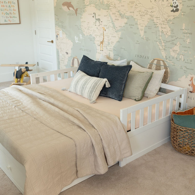 Neutral travel-themed nursery with white toddler bed, beige quilt and decorative pillows, large world map wall mural, wooden toy airplane and woven storage basket