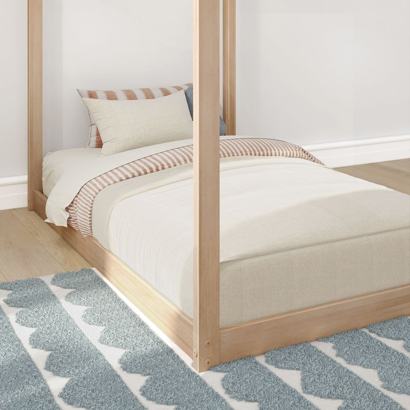 Close-up of light wood four-poster bed with beige bedding, striped pillow and blue textured geometric area rug in minimalist bedroom