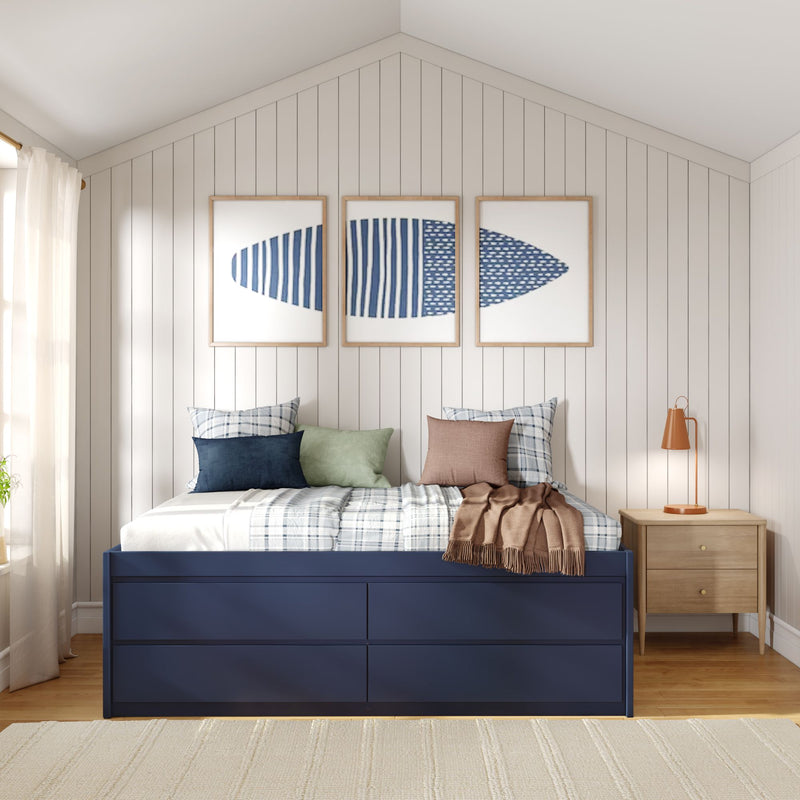 Coastal bedroom with shiplap wall, blue surfboard triptych above navy storage bed, plaid pillows and wood nightstand lamp.