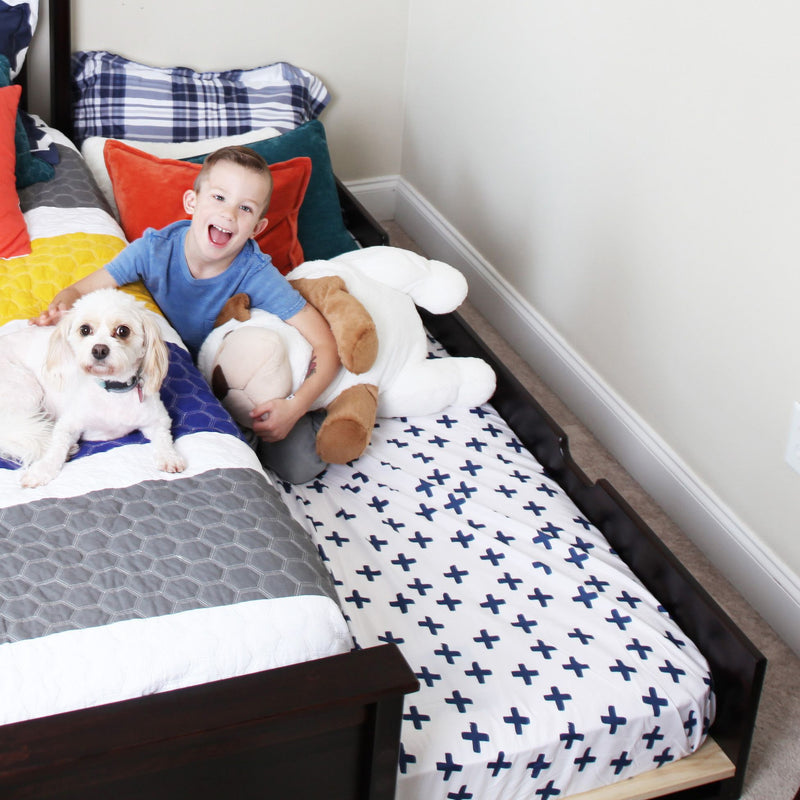 Smiling young boy in a kids' bedroom lying on a bed with a small white dog and stuffed animal on navy cross-patterned sheets and colorful pillows