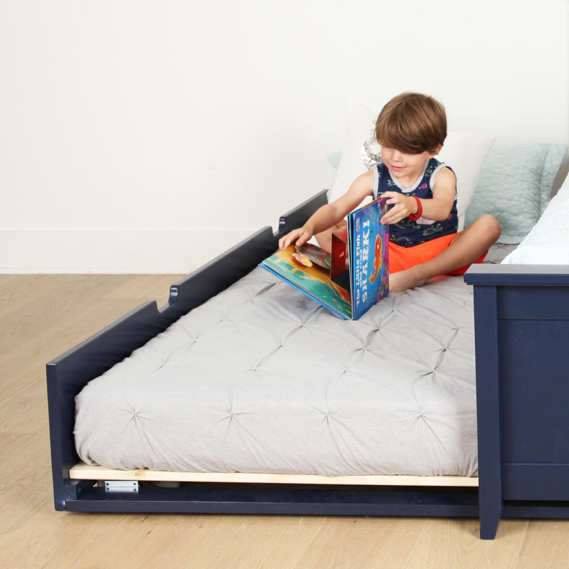 Young child reading a colorful pop-up book on a navy wooden toddler bed with a light gray tufted comforter in a bright minimalist bedroom