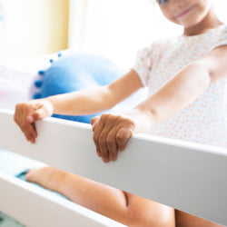 Child holding onto the rails of a white bed frame in a bright bedroom setting.