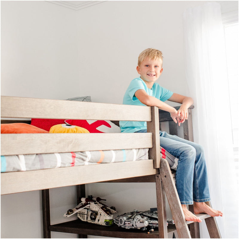 Smiling blond boy sitting on the ladder of a wooden loft bed in a bright modern kids' bedroom with colorful pillows and a toy spaceship underneath