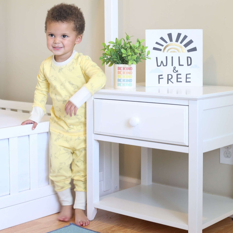 Toddler bedroom decor: toddler in yellow pajamas standing beside white nightstand with "Wild & Free" sign, potted plant, and crib