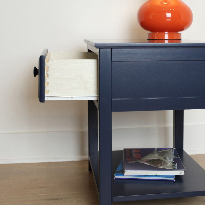 Navy blue nightstand with open drawer, orange table lamp on top and stacked books on lower shelf on hardwood floor