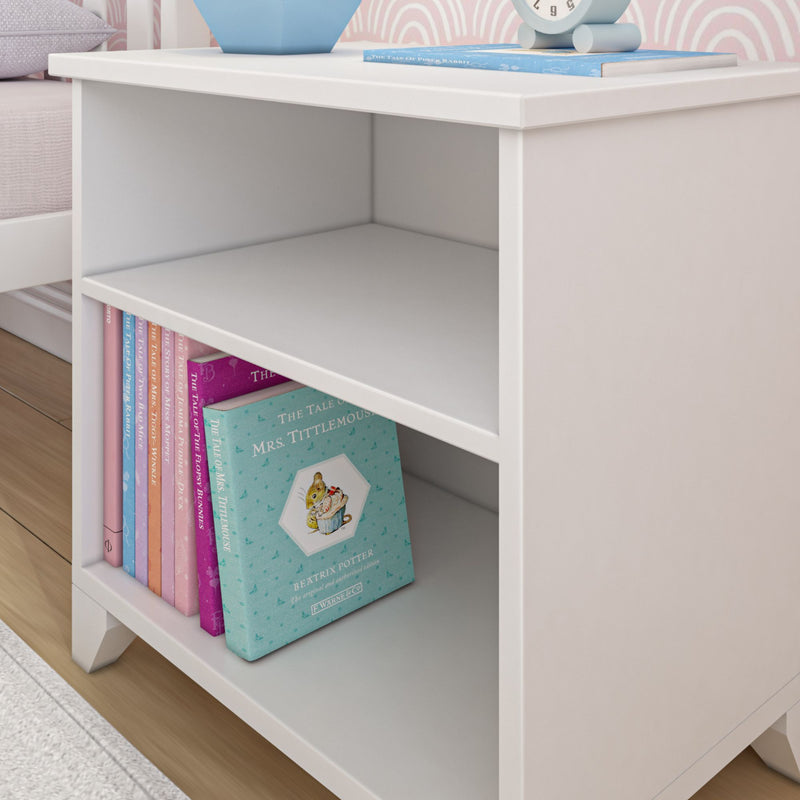White bedside table with cubby shelves holding pastel children's books and a mint alarm clock in a pink nursery