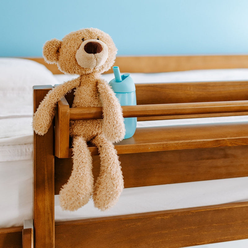 Plush beige teddy bear sitting on a wooden bed rail next to a blue sippy cup in a cozy toddler bedroom nursery