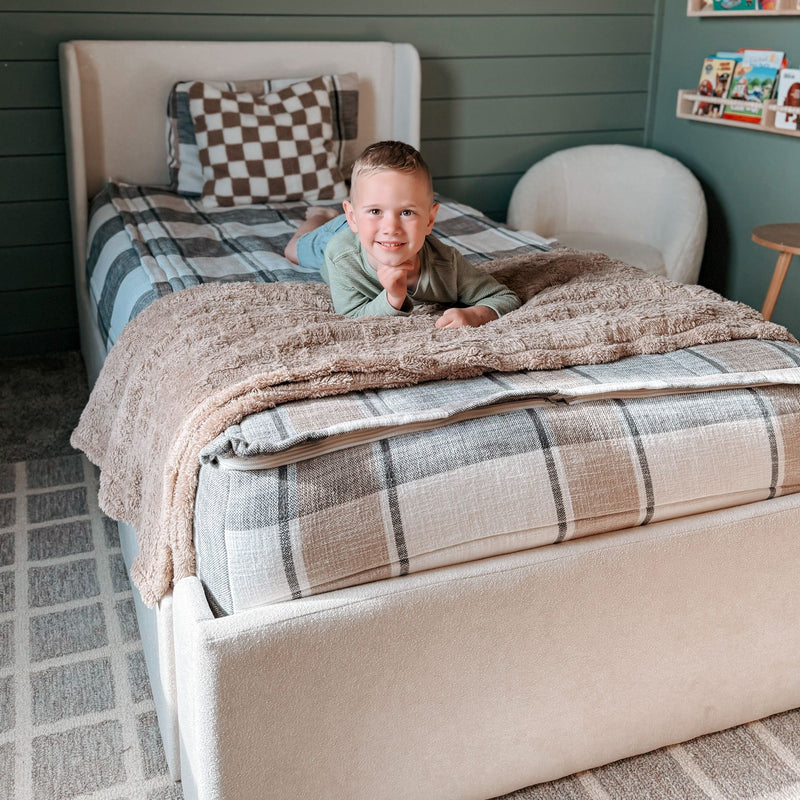Smiling young boy lying on a bed in a cozy kids' bedroom with plaid bedding, beige plush blanket and children's bookshelf