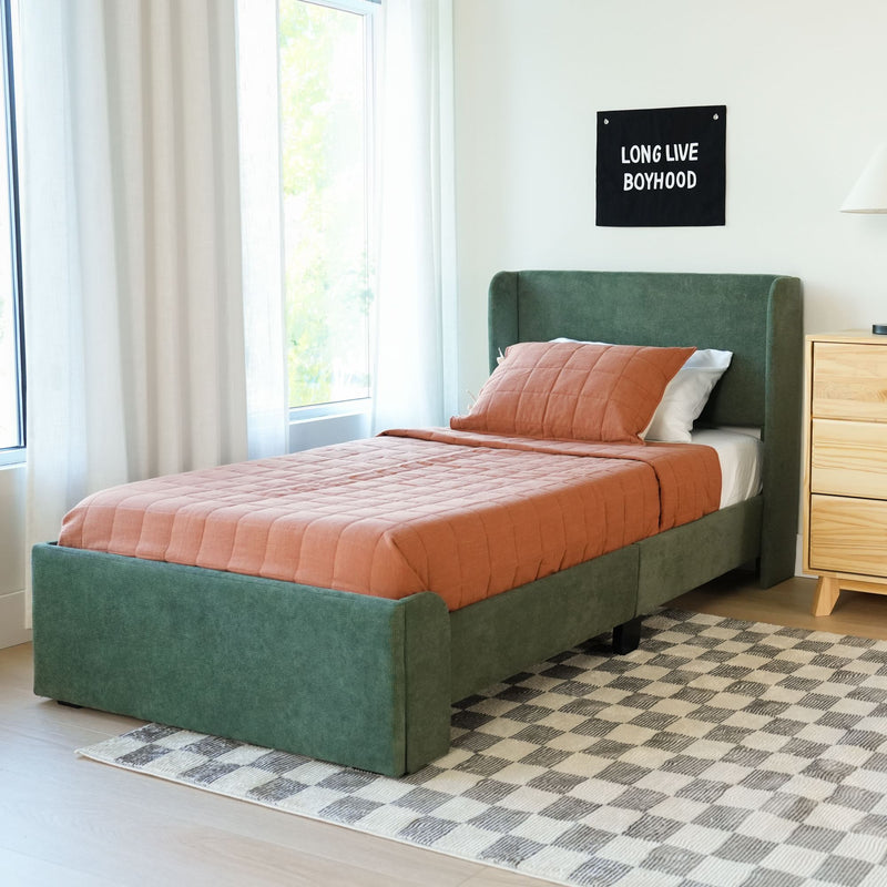 Modern boy's bedroom with green upholstered twin bed, rust quilted bedding, wooden nightstand and lamp, sheer curtains and "Long Live Boyhood" wall hanging