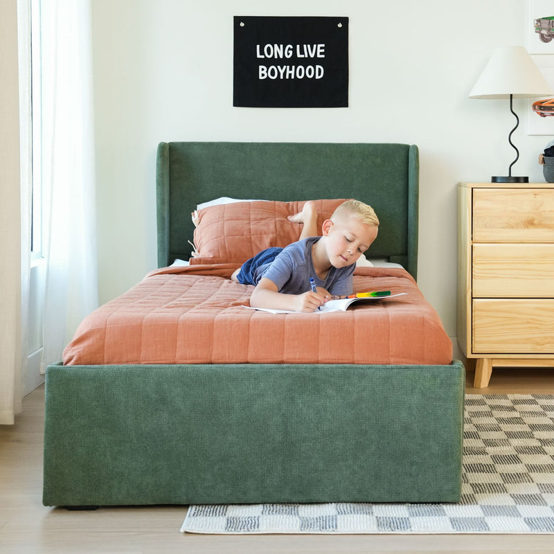 Boy coloring on bed in modern kids bedroom with green upholstered headboard, peach quilt, wooden nightstand and "Long Live Boyhood" wall sign
