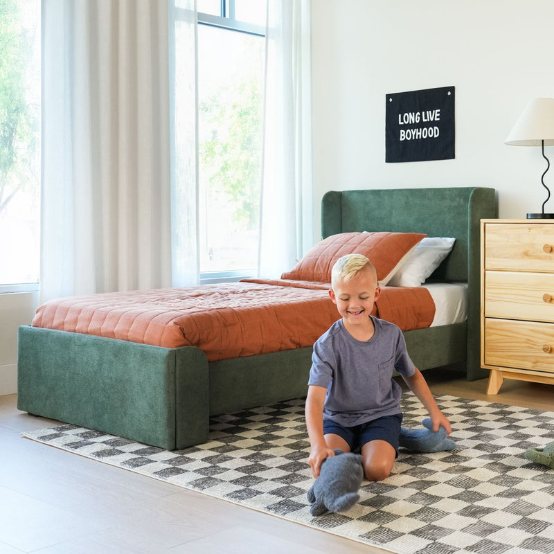 Modern kids bedroom with young boy playing with a stuffed animal on a black-and-white checkered rug, green upholstered bed with rust quilt, wooden nightstand, natural light and "Long Live Boyhood" wall banner