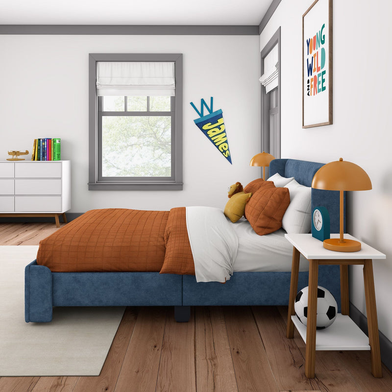 Modern children's bedroom with blue upholstered bed, orange bedding and pillows, bedside table with orange lamp and clock, soccer ball and stuffed lion, white dresser with books and toy plane, framed "Young Wild and Free" wall art and "James" pennant