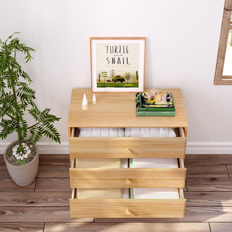 Light wood three-drawer dresser with folded linens in open drawers, framed "Turtle and Snail" art, stacked books and potted plant on hardwood floor