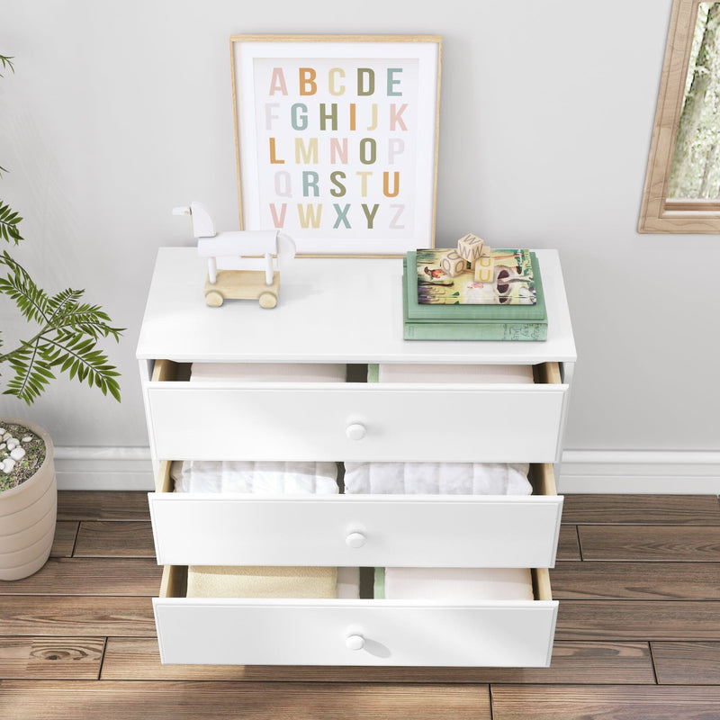 White nursery dresser with three open drawers filled with folded baby clothes, framed alphabet print, wooden pull toy and children's books on top, potted plant and hardwood floor