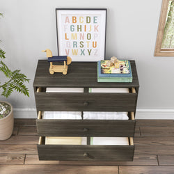 Dark wooden dresser with open drawers showing folded linens, topped with a wooden toy, alphabet art, and children's books, next to a potted plant on hardwood flooring.