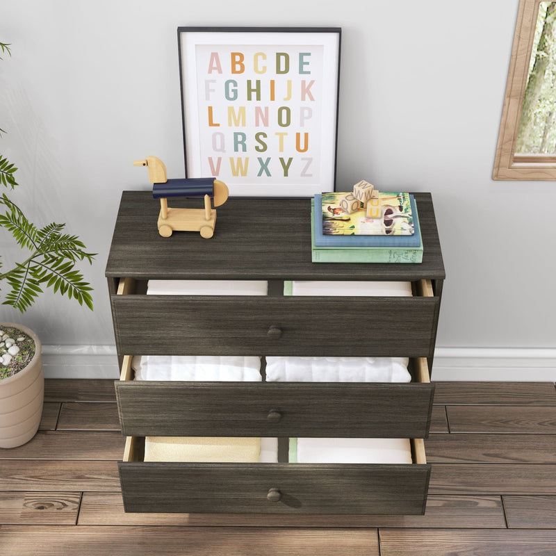Dark wooden dresser with open drawers showing folded linens, topped with a wooden toy, alphabet art, and children's books, next to a potted plant on hardwood flooring.