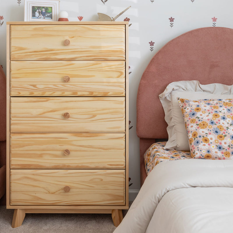 Light pine wooden five-drawer dresser next to blush pink upholstered bed with floral daisy bedding in girls' bedroom