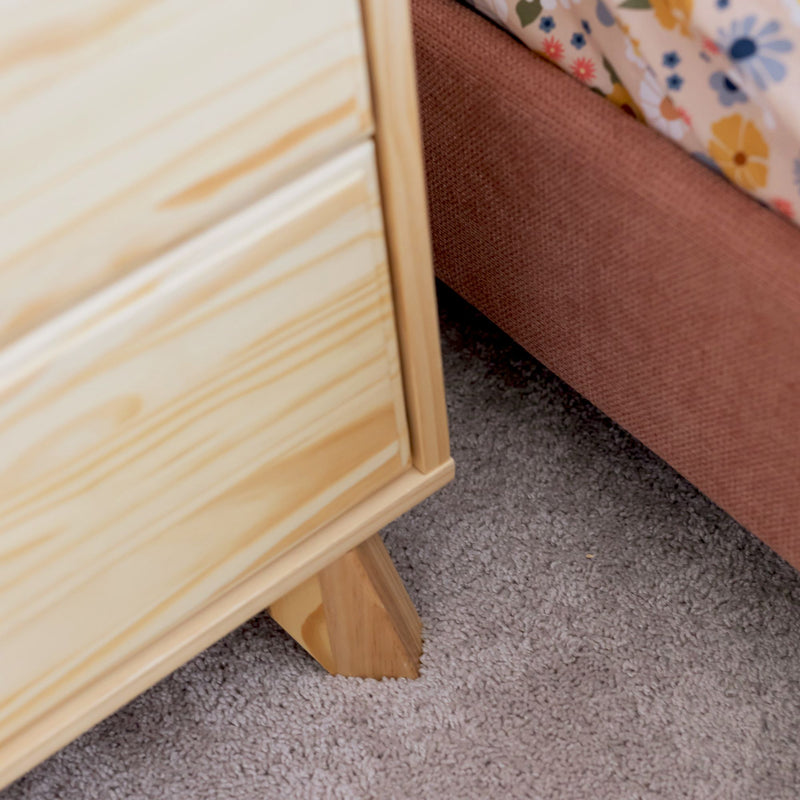 Close-up of light pine wooden nightstand leg on beige carpet beside a pink upholstered bed frame with floral bedding