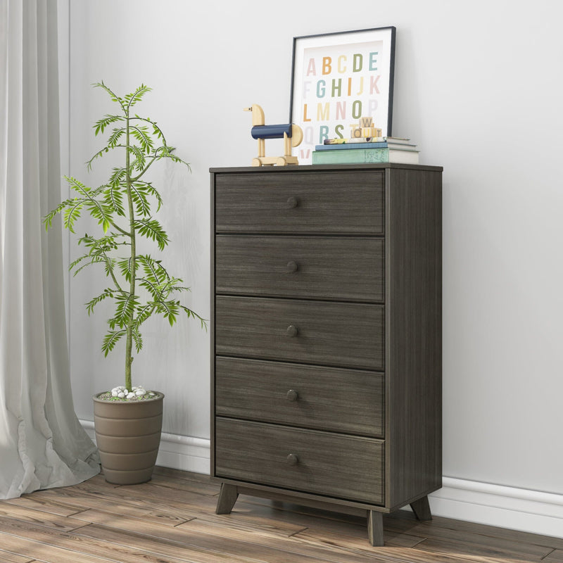 Dark gray wood five-drawer tall dresser with round knobs and angled legs in a modern bedroom setting with potted plant and framed alphabet art