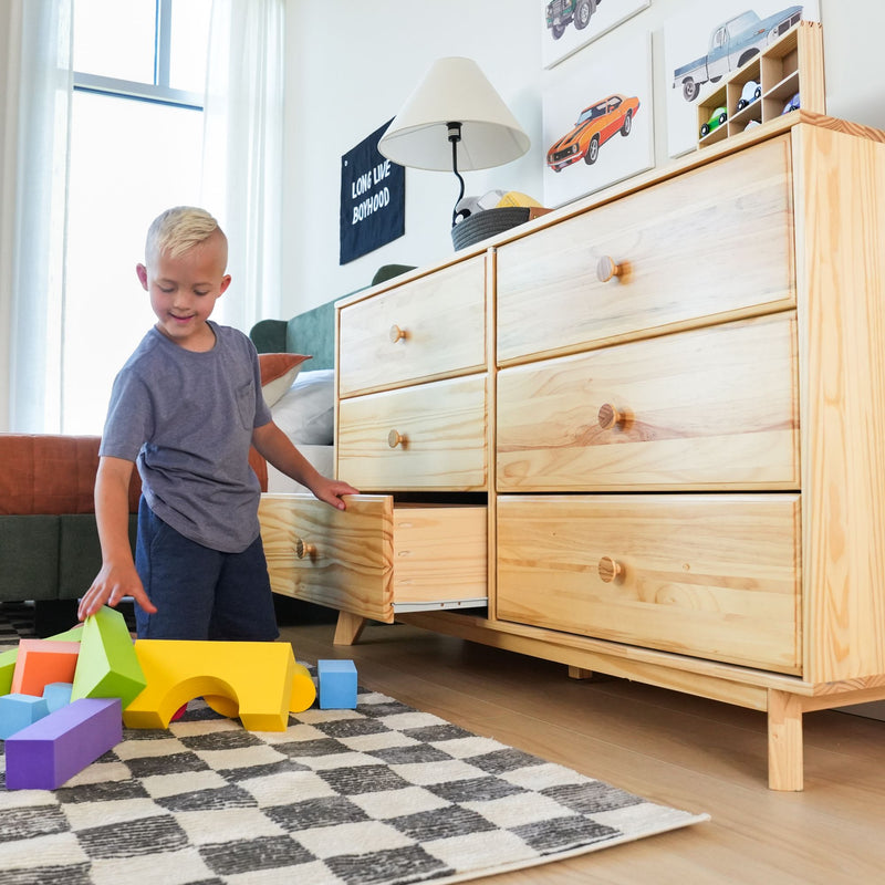 Young boy playing with colorful foam blocks on a rug in a bright modern kids' bedroom beside a light wood dresser with open drawers and car wall art