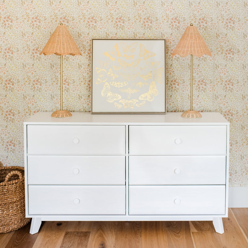 White six-drawer dresser with rattan table lamps, framed gold butterfly art, woven basket on wood floor and floral wallpaper backdrop