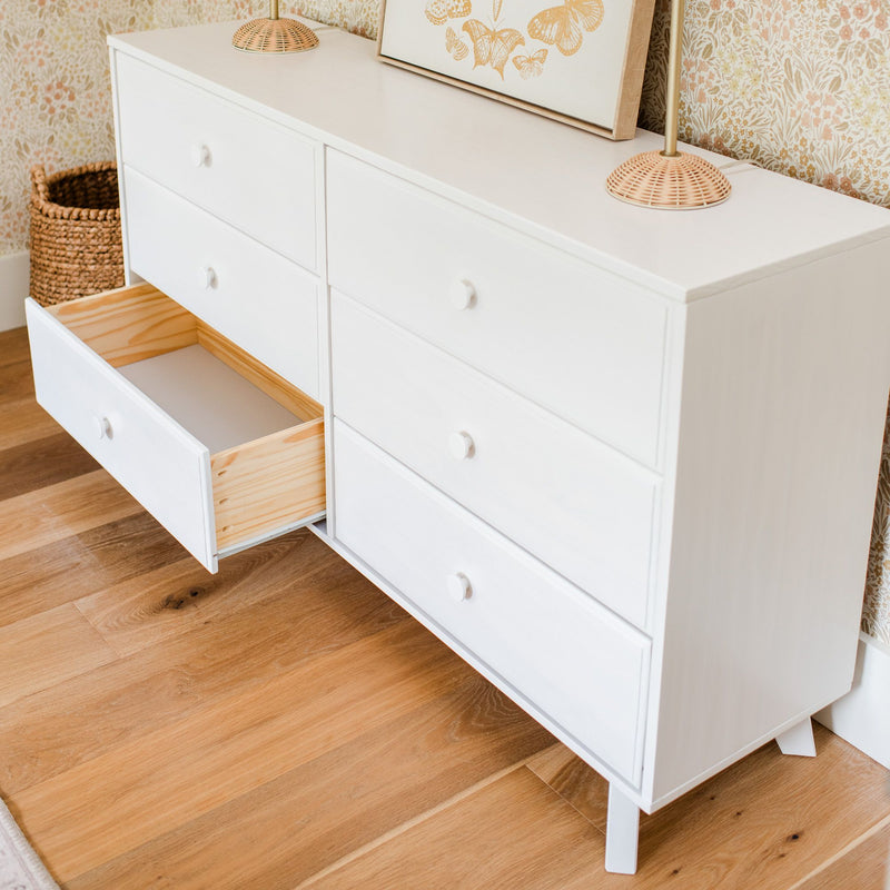 Modern white six-drawer dresser with one open drawer on oak floor, styled with rattan lamp, woven basket and floral wallpaper