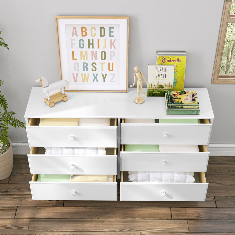 White six-drawer nursery dresser with open drawers showing folded linens, wooden toys, alphabet wall art and children's books on hardwood floor