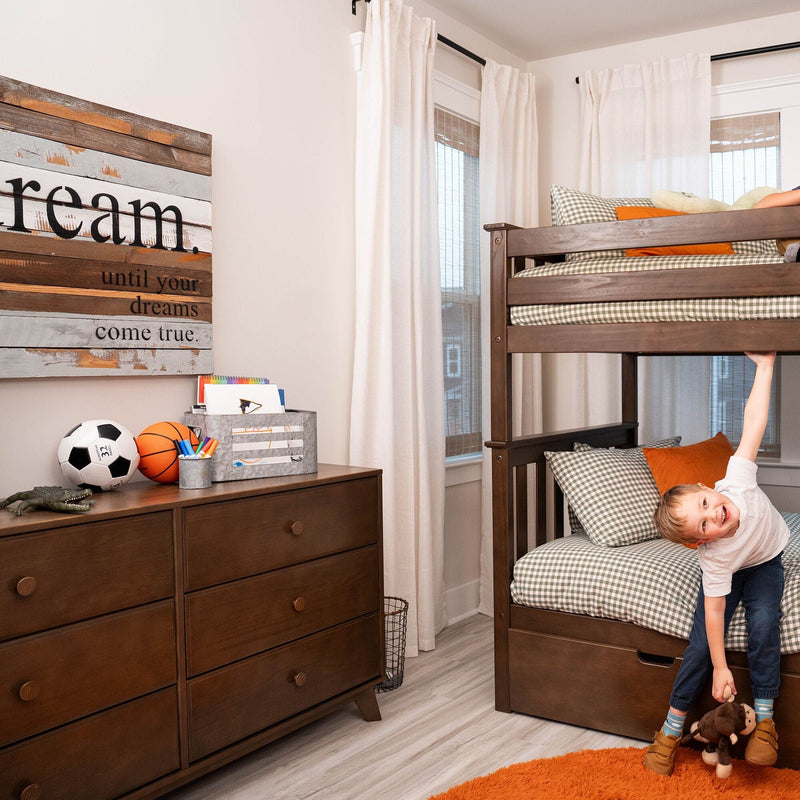 Young boy in modern kids bedroom with dark wooden bunk bed, checkered bedding, wooden dresser with soccer and basketball, orange accents and "dream" wall art