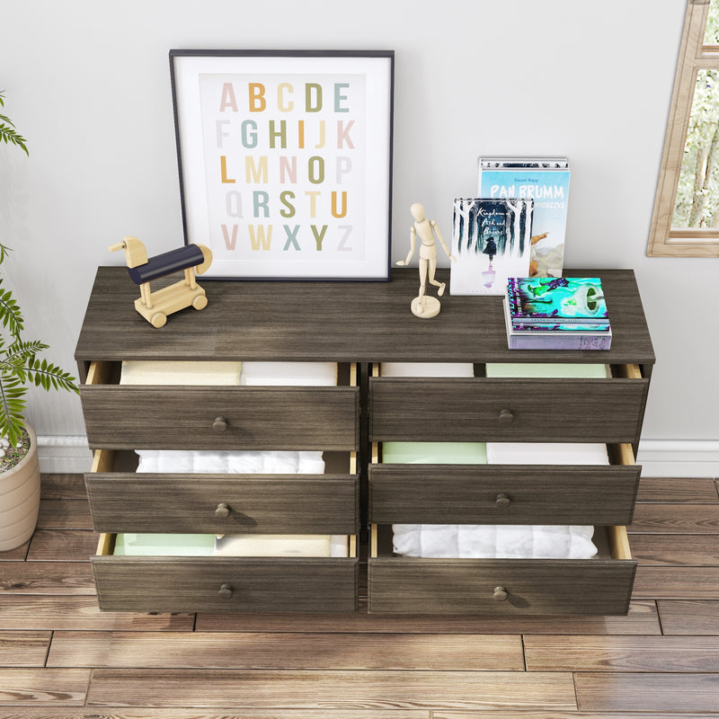 Dark wood six-drawer dresser with several drawers open showing folded baby blankets, framed pastel alphabet print, wooden pull toy, artist mannequin and stacked books on hardwood floor