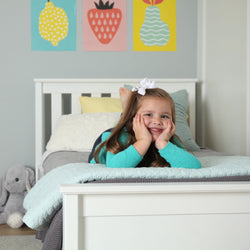 Smiling young girl lying on a white bed in a pastel kids' bedroom with fruit wall art, mint quilt and gray stuffed bunny.