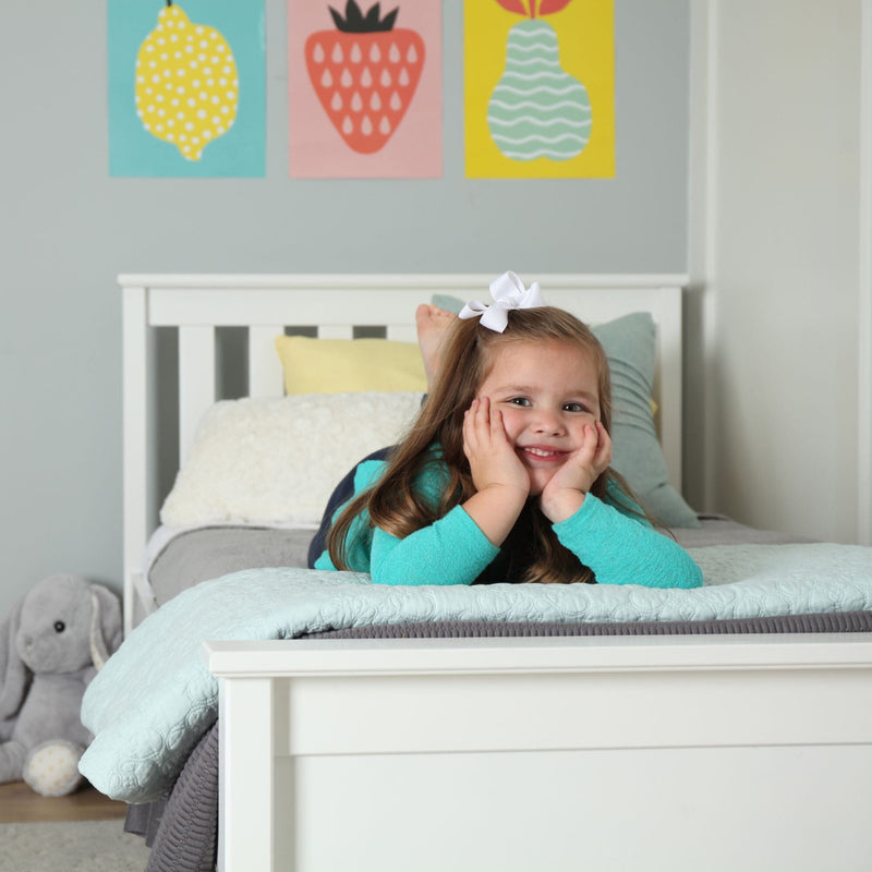 Smiling young girl lying on a white bed in a pastel kids' bedroom with fruit wall art, mint quilt and gray stuffed bunny.