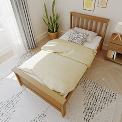 Scandinavian-style bedroom with wooden single bed, yellow duvet, spotted accent pillow, bedside lamp, potted snake plant, and sunlight.