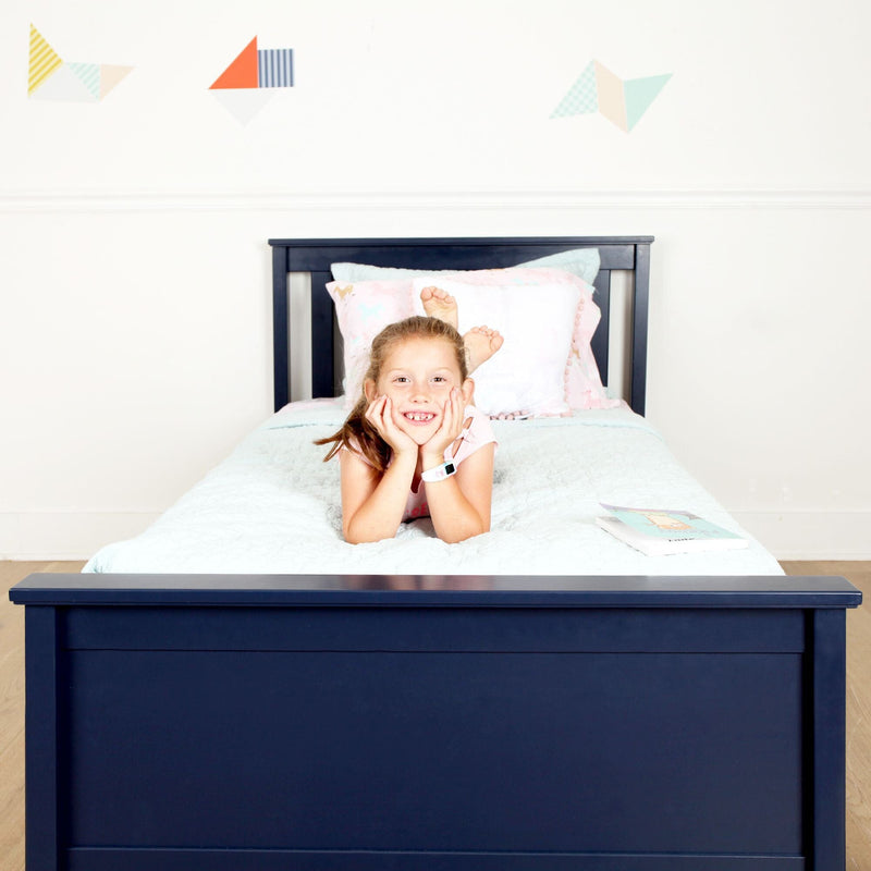 Smiling girl lying on a navy blue wooden twin bed with white quilt, pastel pillows and a children's book in a bright bedroom